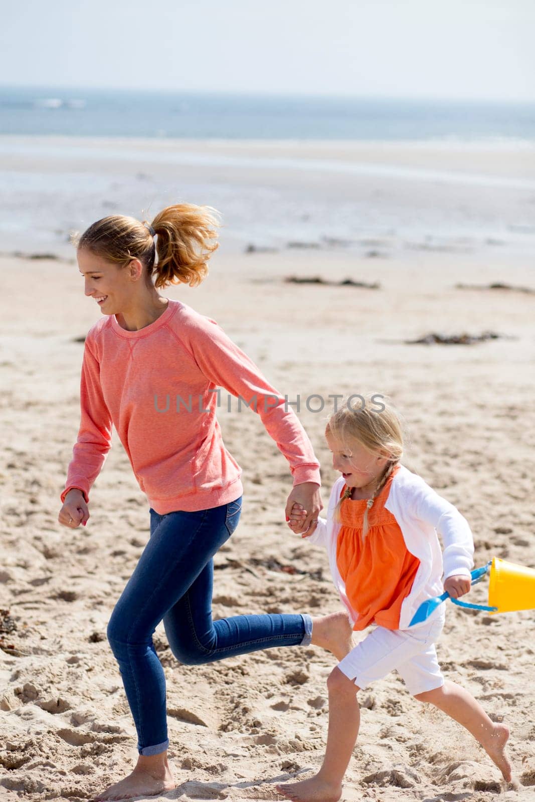 Young mother and daughter running along the beach with a bucket and spade. They are both laughing and smiling.