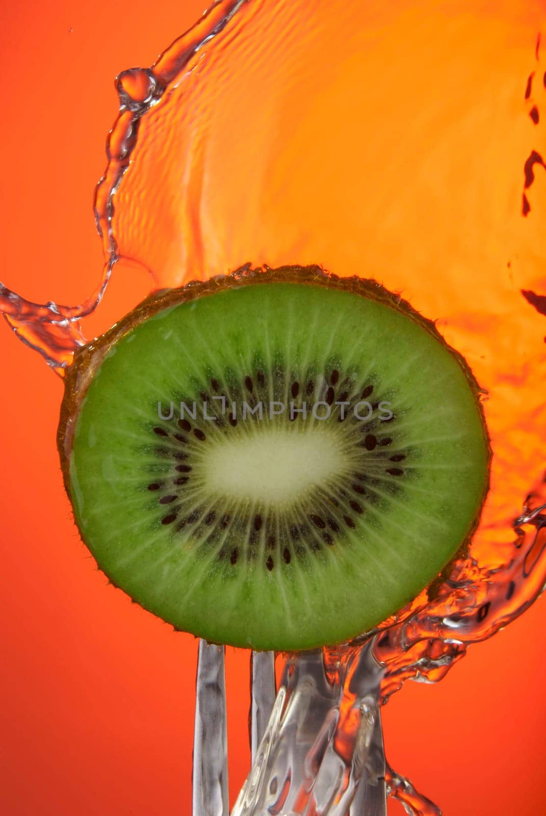 Close up of kiwi fruit center on fork with water splash and orange color background, concept photo. Ideal for healthy eating campaign or fitness poster.