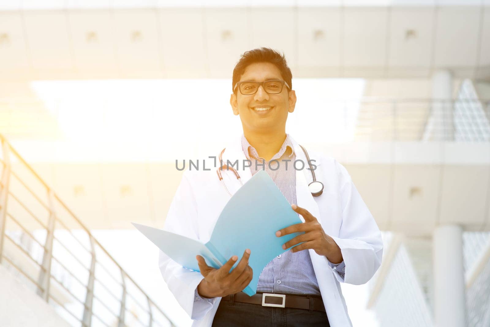 Portrait of Asian Indian medical doctor smiling and reading on medical report, standing outside hospital building, beautiful golden sunlight at background.