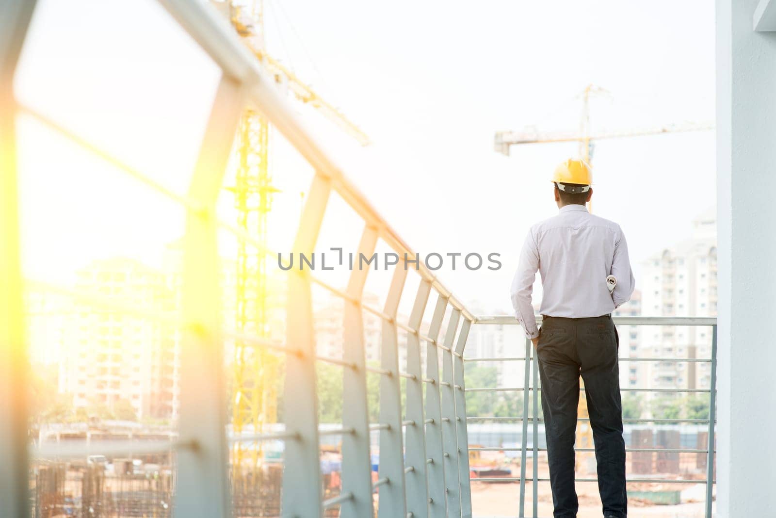 Rear view Asian Indian male site contractor engineer with hard hat holding blue print paper inspecting at construction site, crane with golden sunlight at the background.