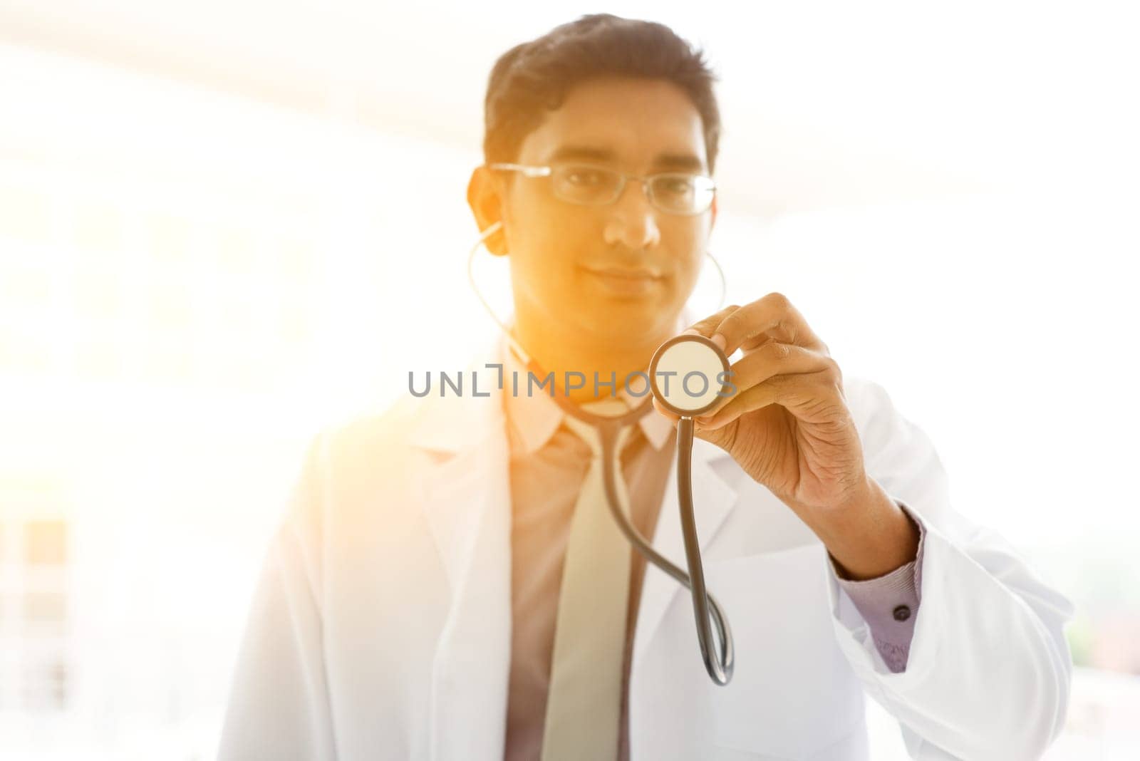 Portrait of Asian Indian medical doctor holding stethoscope standing outside hospital building, focus on stethoscope , beautiful golden sunlight at background.