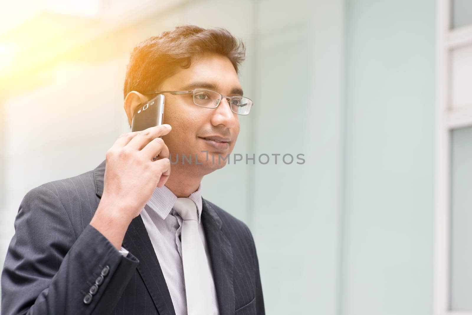 Portrait of Asian Indian business man using smartphone, outside modern office building block, beautiful golden sunlight at background.