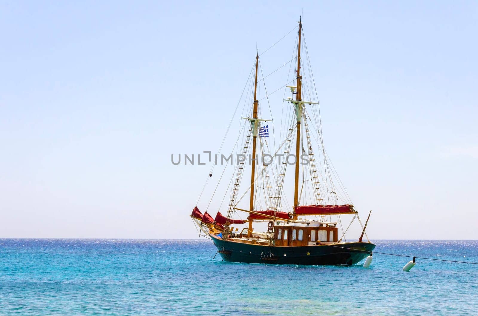 A traditional wooden sail boat anchored on the coast of Mykonos, Greece. The wind sails are down and it is tied on the rocks by a rope. A very typical greek island summer holiday scene on crystal clear blue sea.