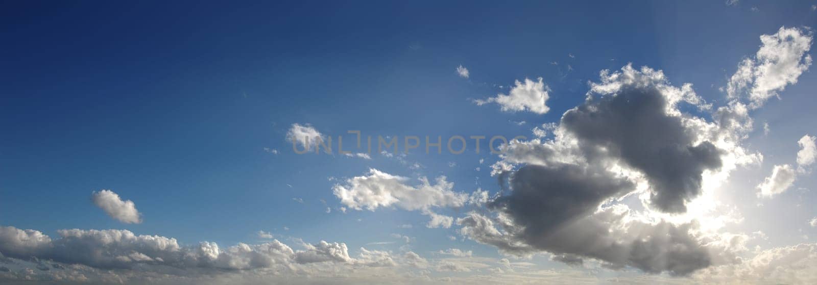 Blue sky with several cumulus clouds in background and a large cloud masking the sun, in the foreground, generating some godrays. Cinemascope format.