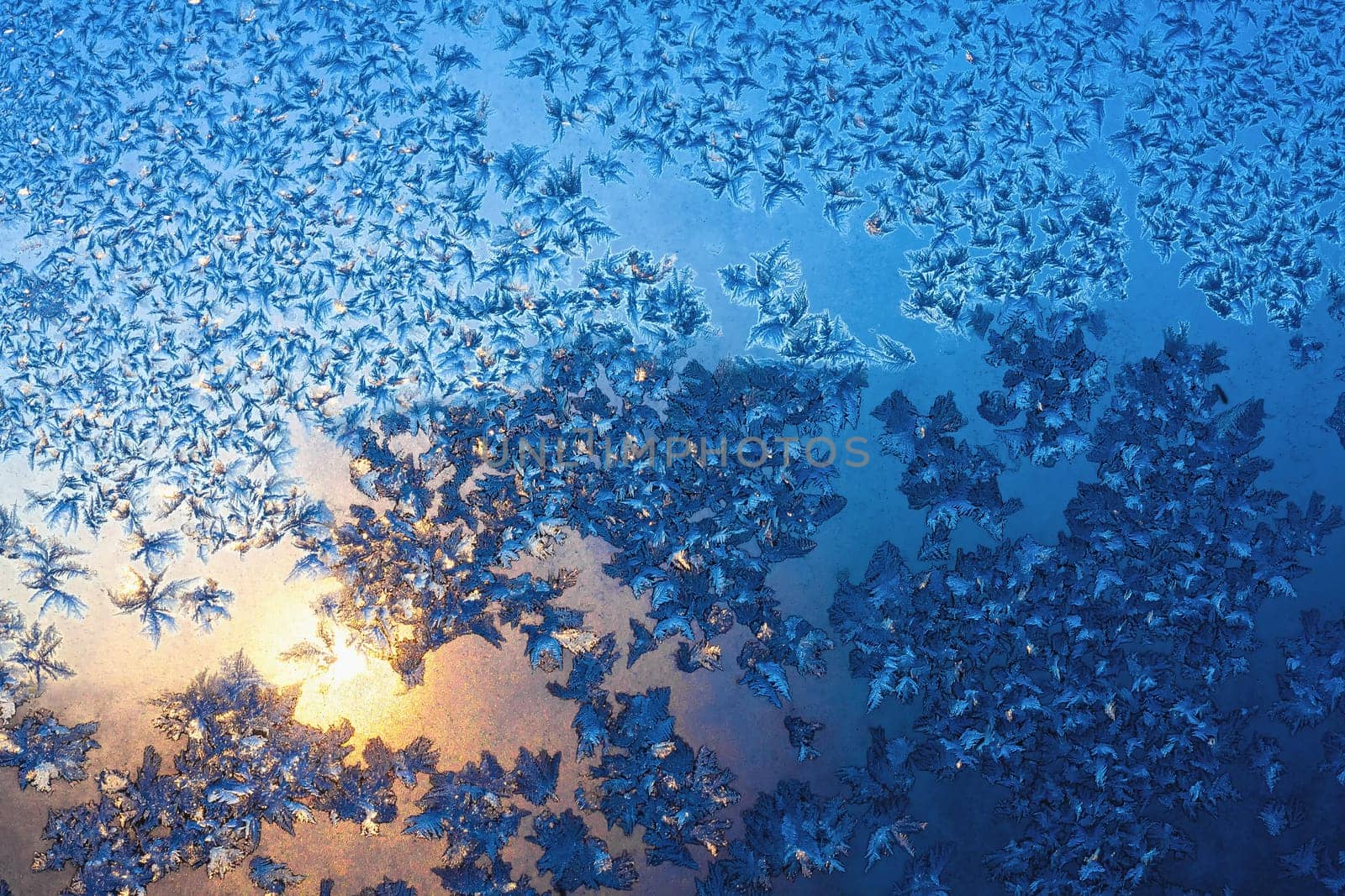 Frozen snowflakes and frost over the sun on frozen Christmas winter window at sunset. Colored in blue tone. Selective focus at center with blur edges of image