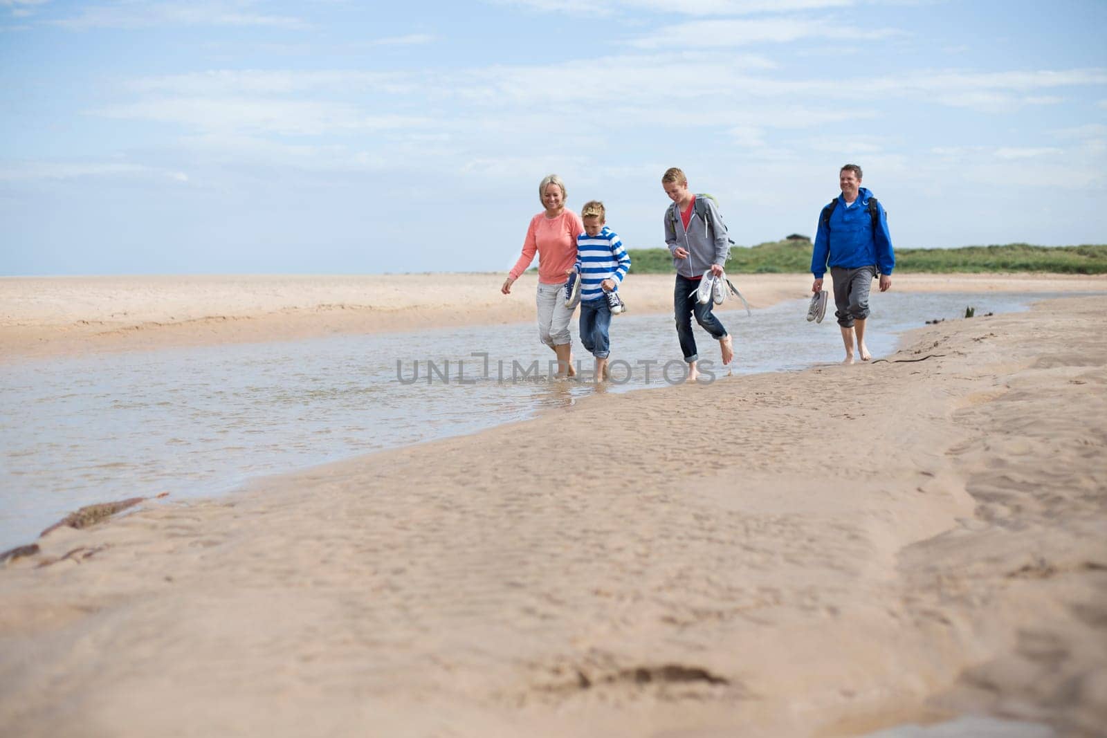 Family of four walking through water and sand at the beach. They are barefoot and wearing warm and casual clothing.