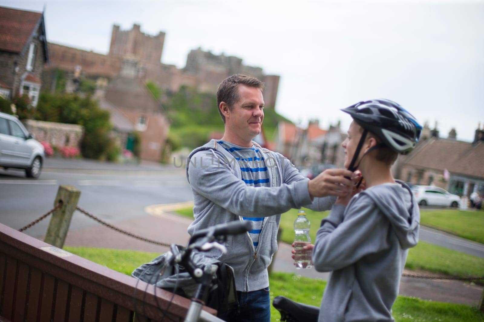 Father putting his helmet on for his son. They are wearing casual clothing and a background of a village and castle can be seen in the distance.