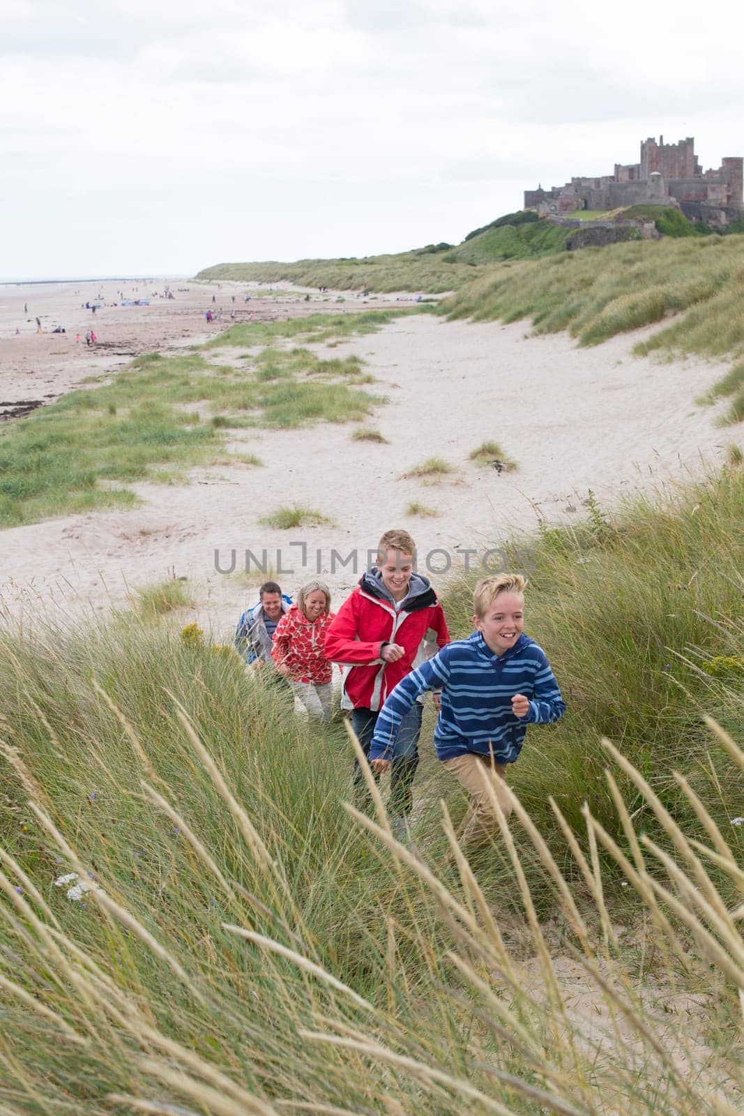 Family of four running up the sand dunes at the beach. They are wearing warm casual clothing and a castle can be seen in the background.