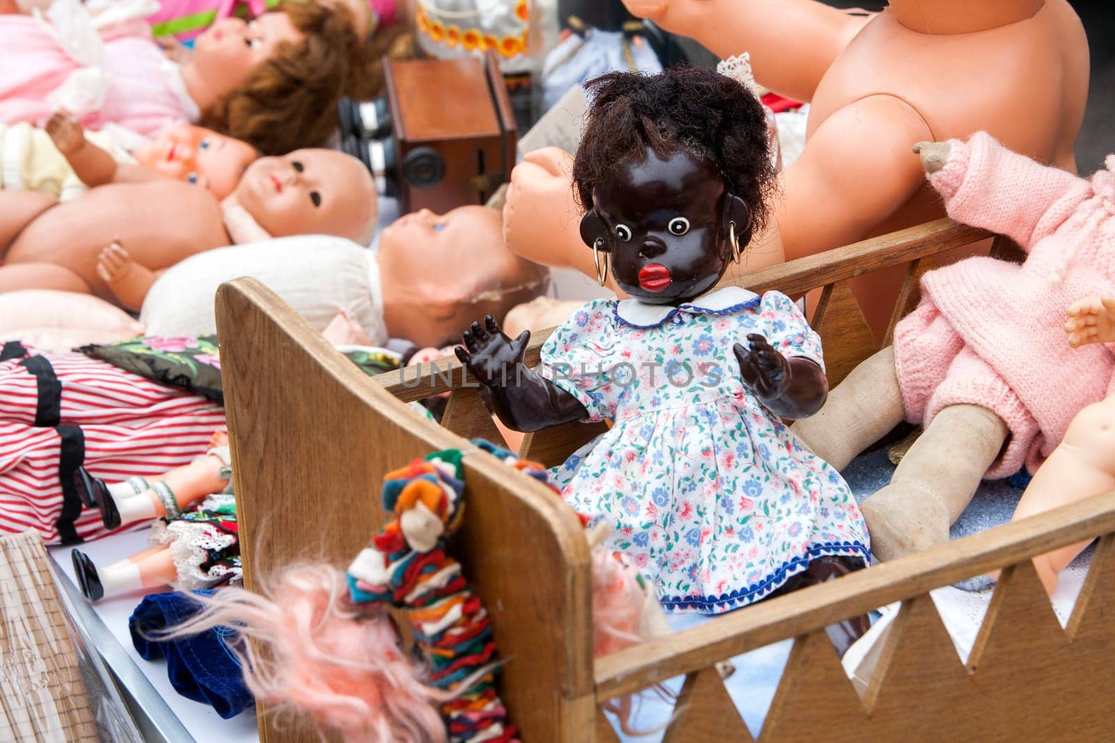 Second hand dolls at a jumble sale in France. Black doll with golden earrings in a wooden crib on the foreground and some white dolls in the background.