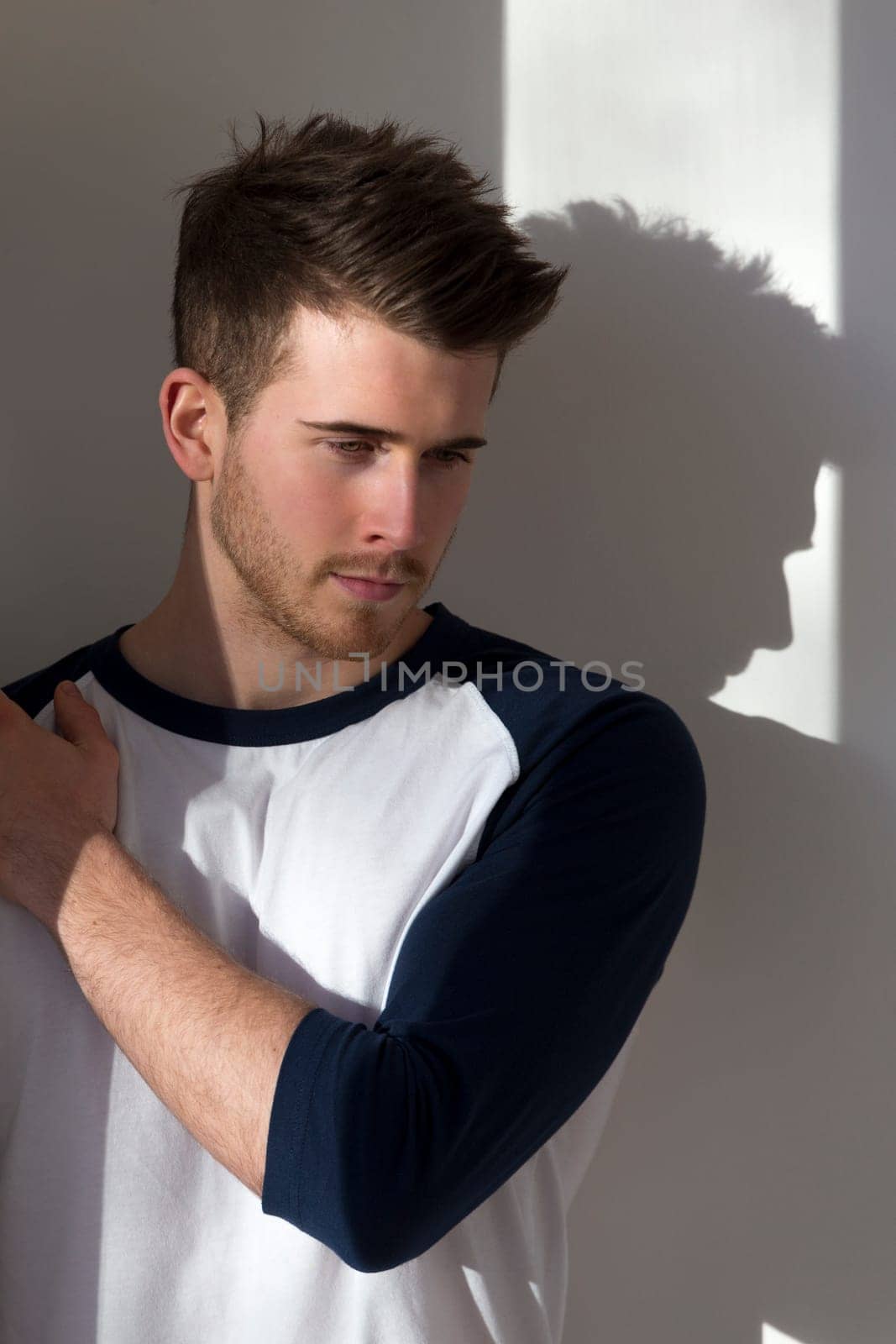 Portrait of an attractive young man, standing against a white wall with sunlight from the window shining on him.
