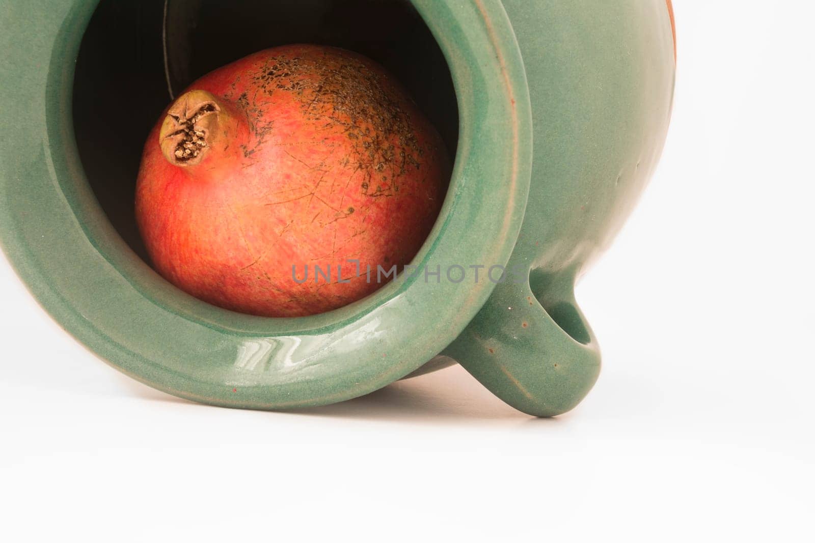 close up view of a red pomegranate inside the greenish earthen jar isolated on white background.