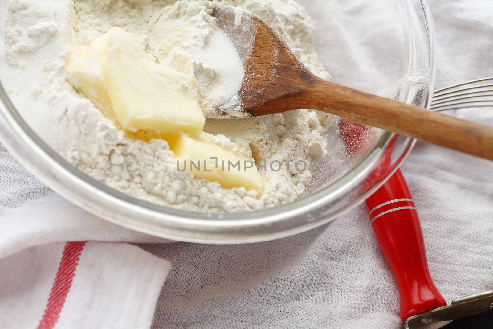 Glass mixing bowl with wooden spoon, butter and flour with pastry blender alongside on dish cloth
