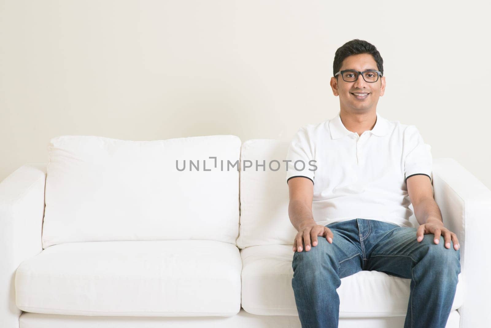 Young single Indian guy sitting lonely on sofa and smiling. Lifestyle Asian man at home. Handsome male model.