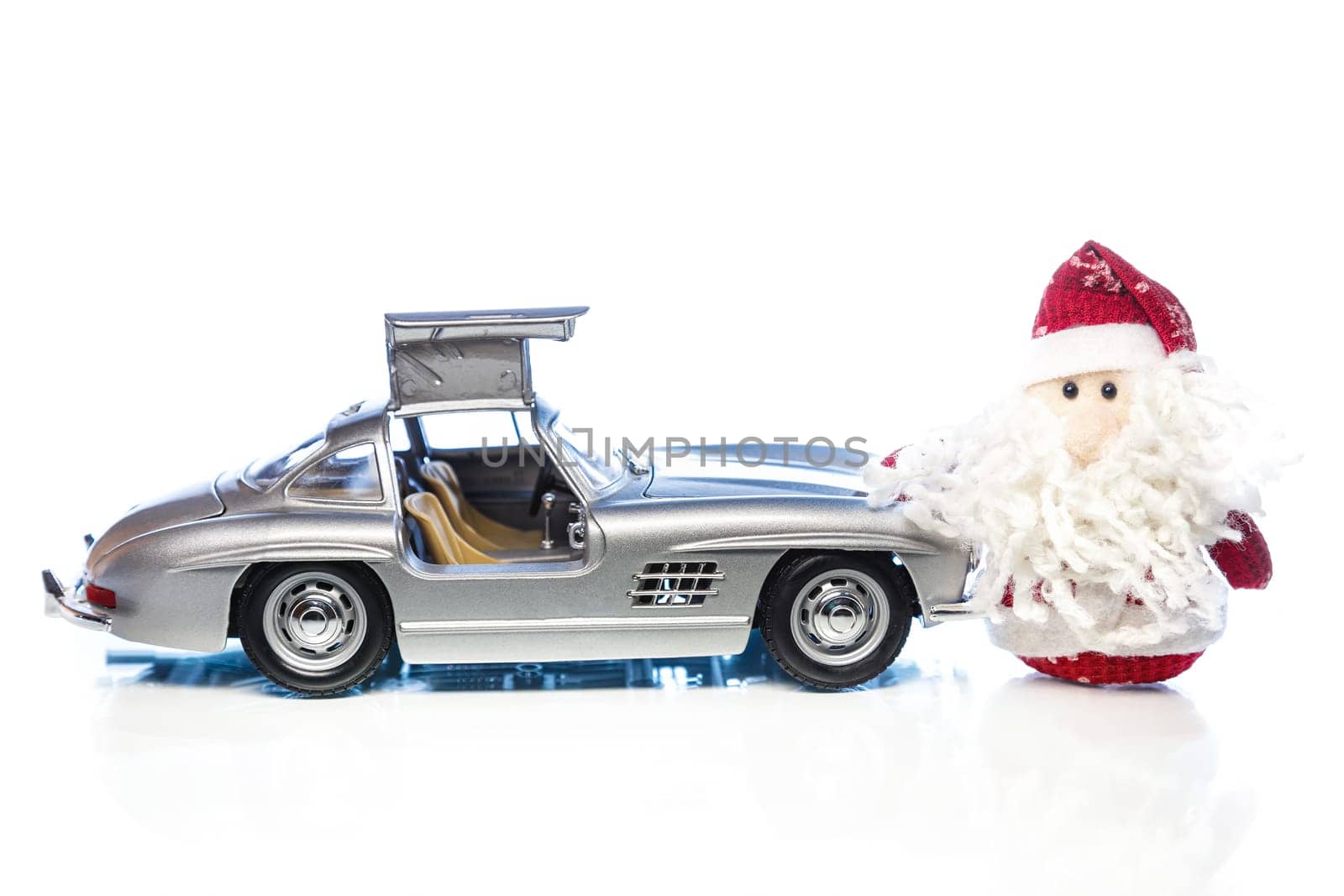 Santa Claus or Father Frost with old vintage automobile on white background with reflection. Main focus of image on Santa Claus and selective on the side turned and opened doors toy retro car