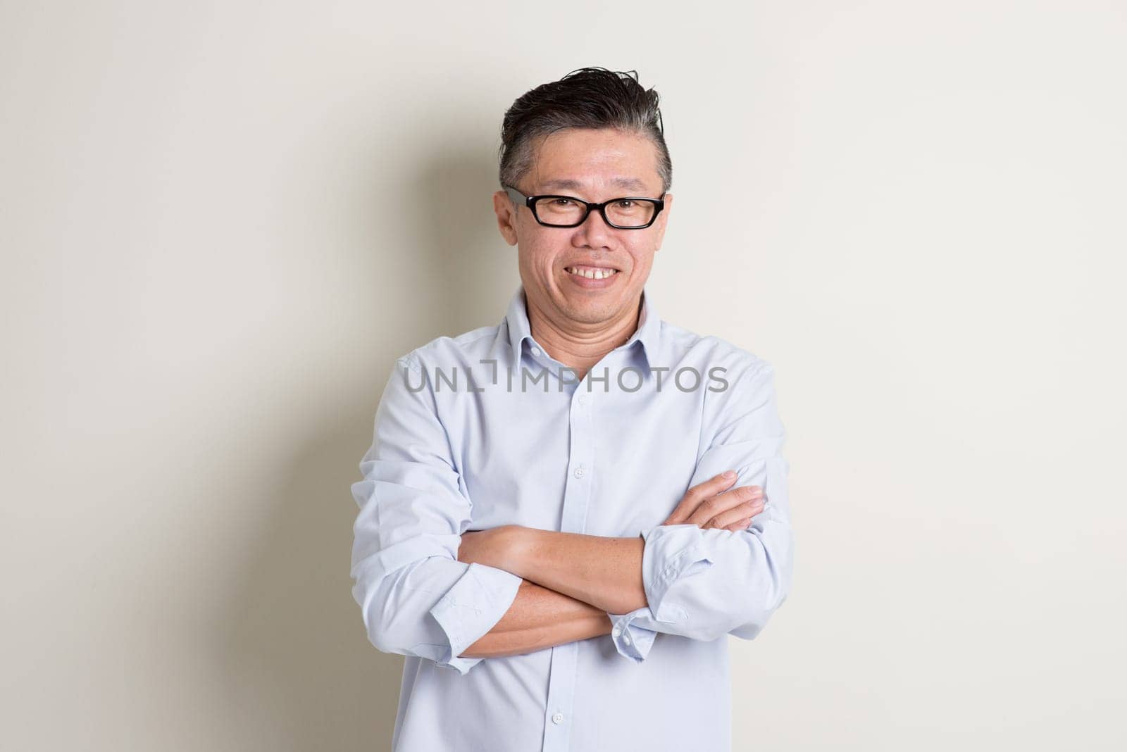 Portrait of confident single mature 50s Asian man in casual business arms crossed and standing over plain background with shadow.