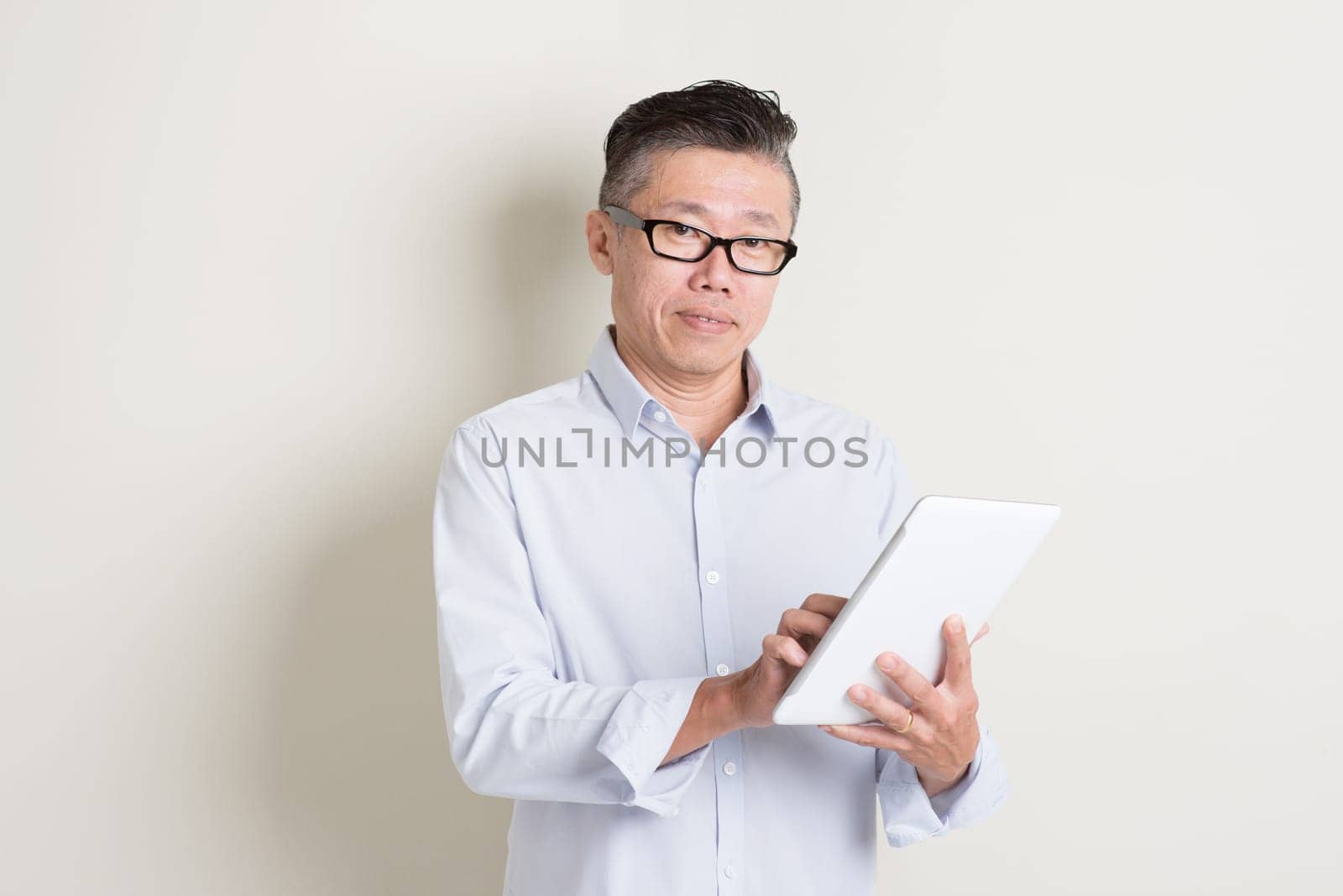 Portrait of single mature 50s Asian man in casual business using digital tablet pc, standing over plain background with shadow. Chinese senior male people.