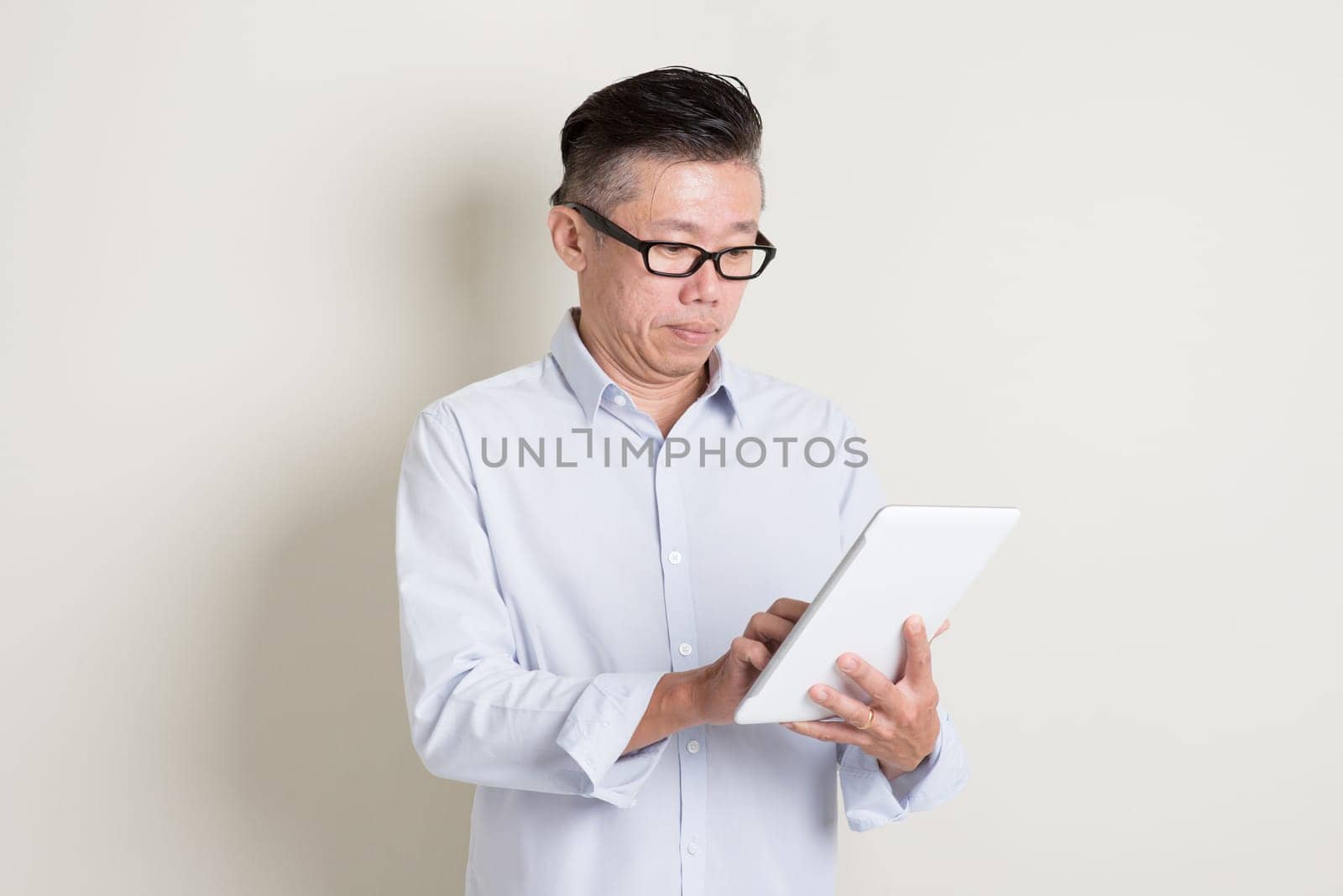 Portrait of single mature 50s Asian man in casual business using digital tablet computer, standing over plain background with shadow. Chinese senior male people.