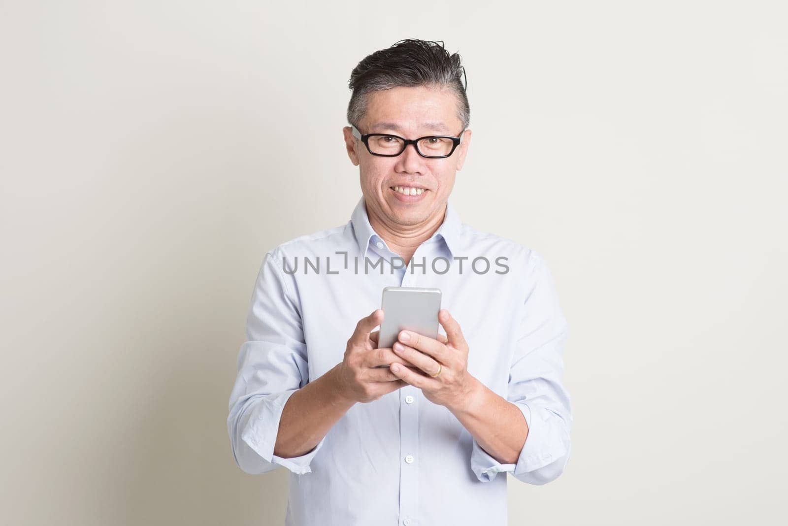 Portrait of single mature 50s Asian man in casual business using mobile applications on smartphone and smiling, standing over plain background with shadow. Chinese senior male people.
