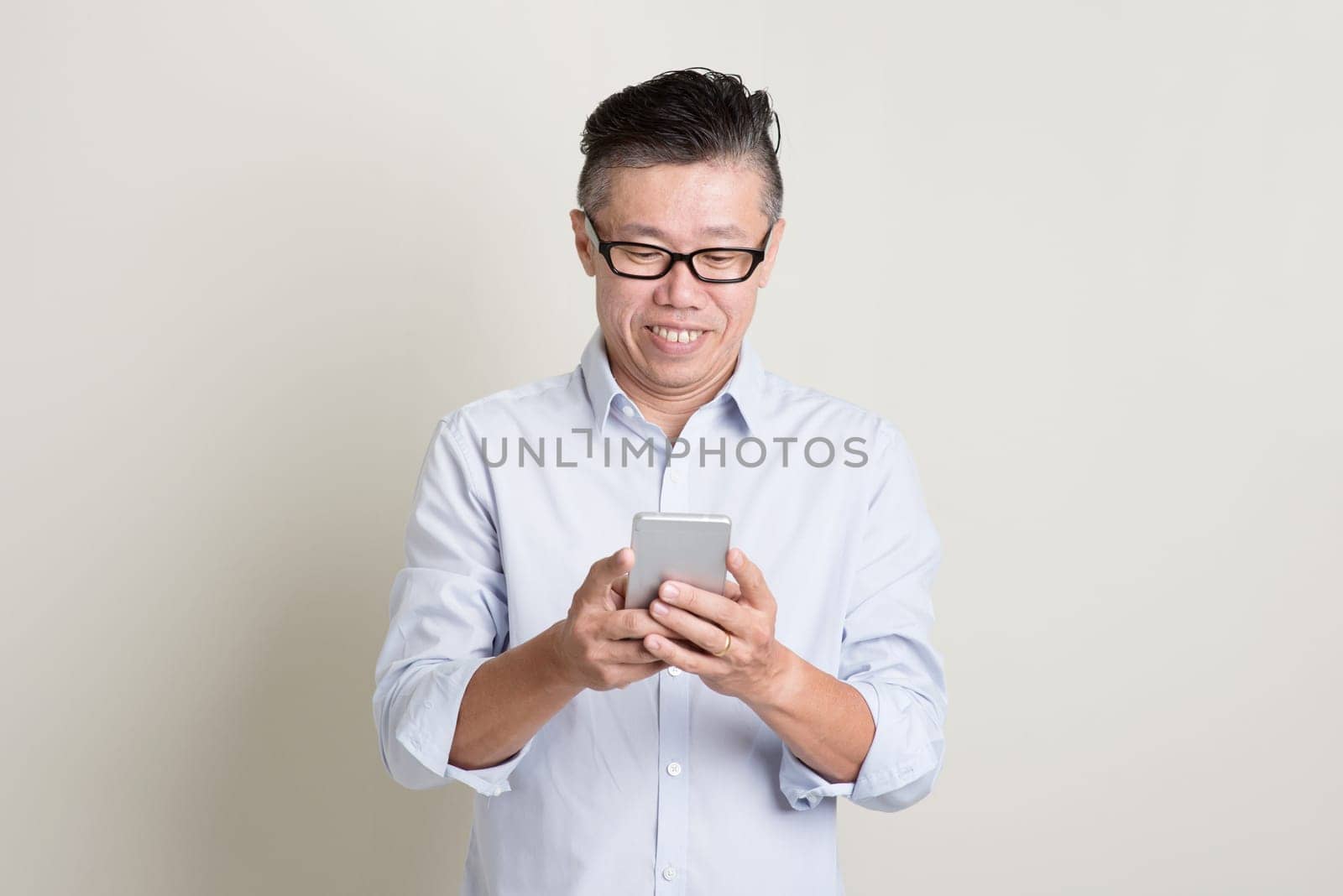 Portrait of single mature 50s Asian man in casual business using mobile apps on smartphone and smiling, standing over plain background with shadow. Chinese senior male people.