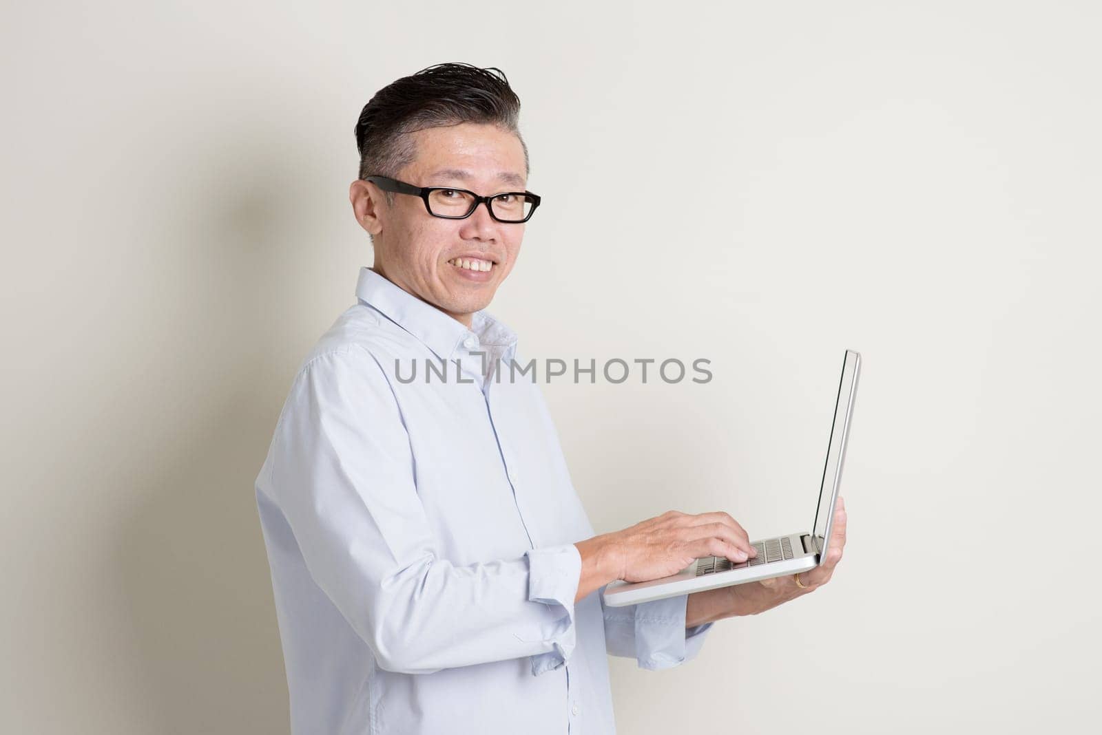 Portrait of single mature 50s Asian man in casual business using notebook pc and smiling, standing over plain background with shadow. Chinese senior male people.