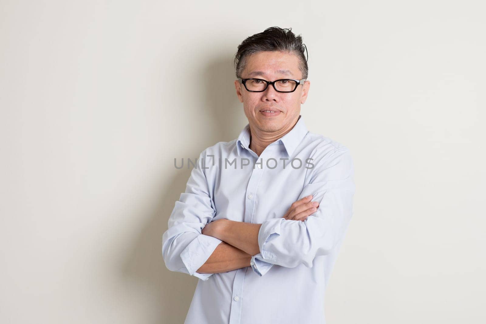 Portrait of single mature 50s Asian man in casual business arms crossed smiling and standing over plain background with shadow.