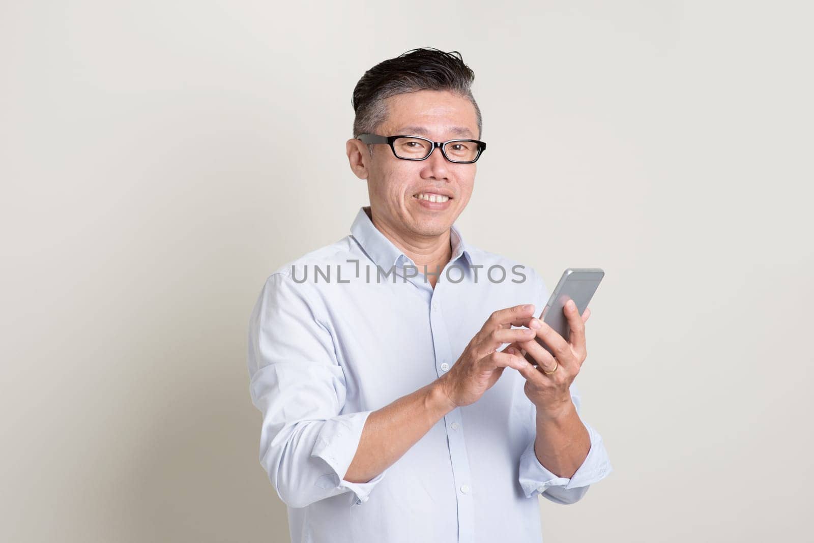 Portrait of single mature 50s Asian man in casual business texting using smartphone and smiling, standing over plain background with shadow. Chinese senior male people.