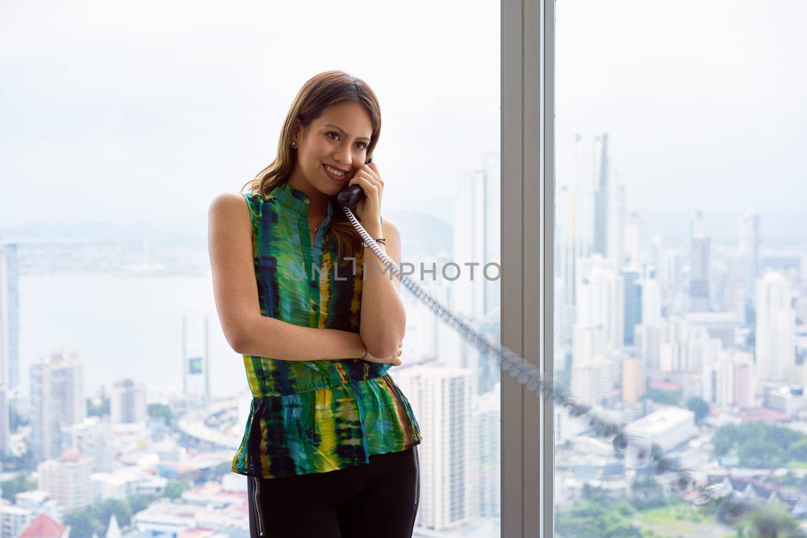 Young Hispanic woman leaning on table in a modern office building, with a beautiful sight of the city in background. She holds a wired telephone with long wire and talks looking away. Copy space