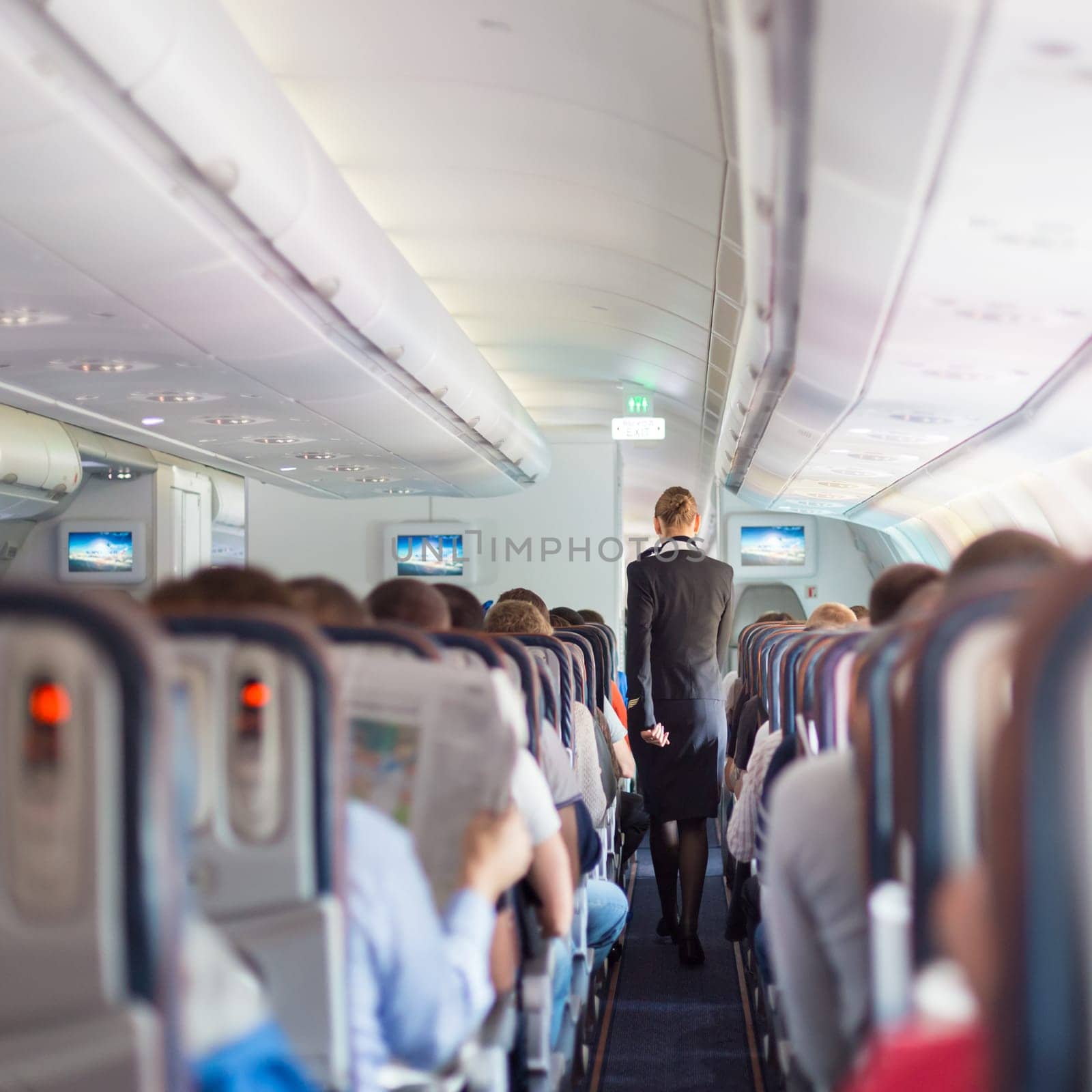 Interior of commercial airplane with passengers on seats during flight. Stewardess in dark blue uniform walking the aisle. Square composition.