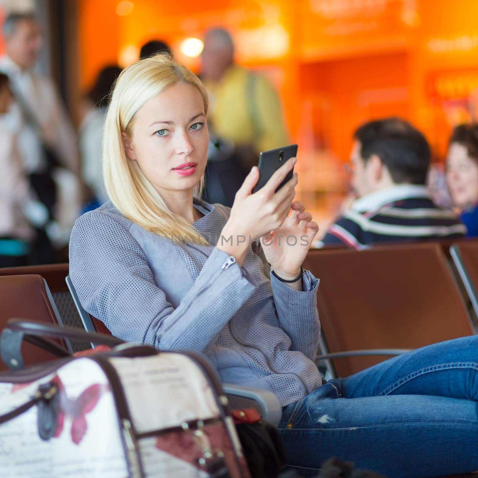 Casual blond young woman using her cell phone while waiting to board a plane at the departure gates. Wireless network hotspot enabling people to access internet conection. Public transport.