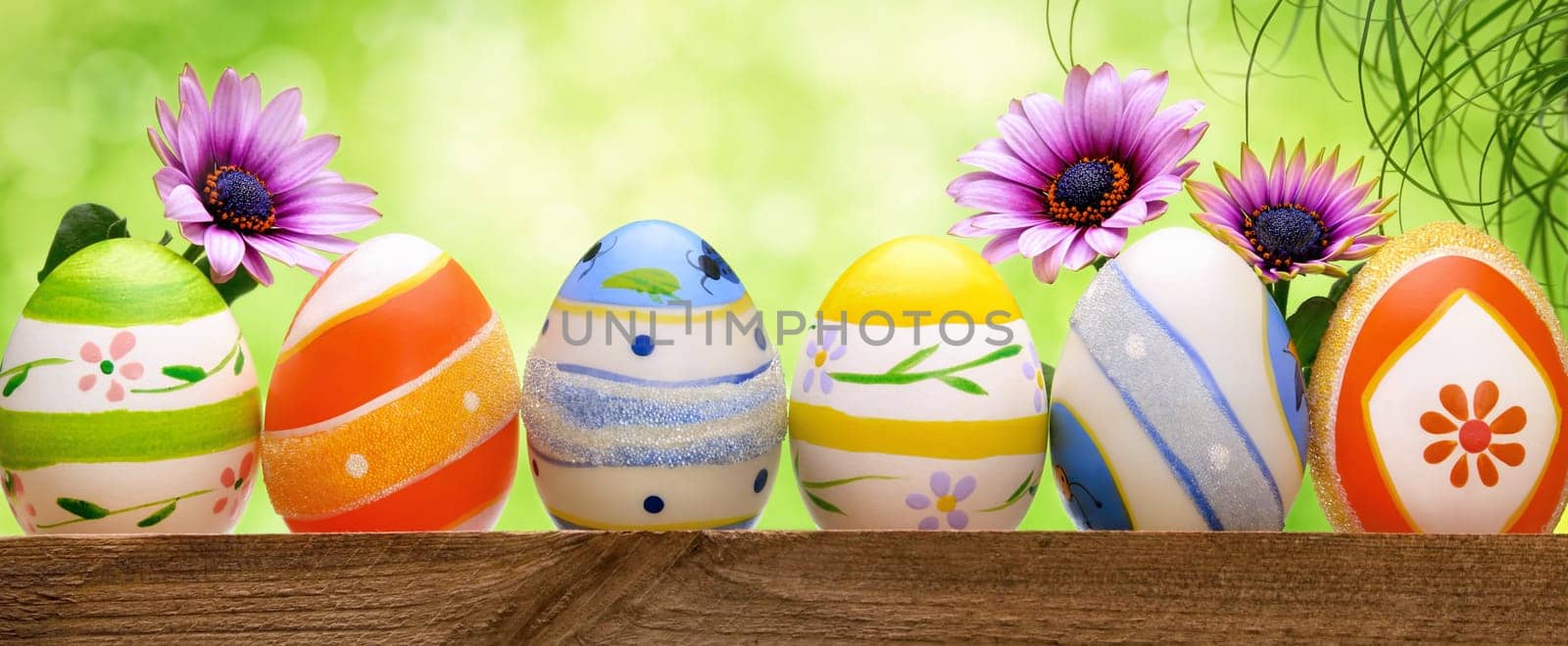 Colorful Easter eggs in a row,decorated with spring flowers and long grass,with bright green bokeh background,panorama format
