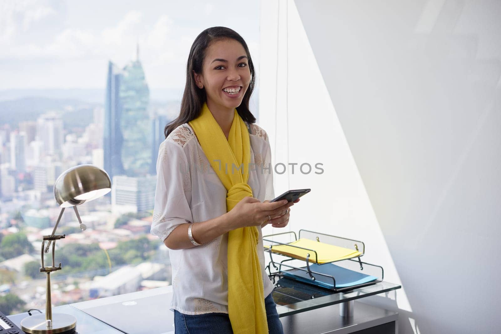 Young multiethnic Chinese Hispanic woman leaning on table in a modern office building, with a beautiful sight of the city in background. She holds a mobile phone and smiles at camera