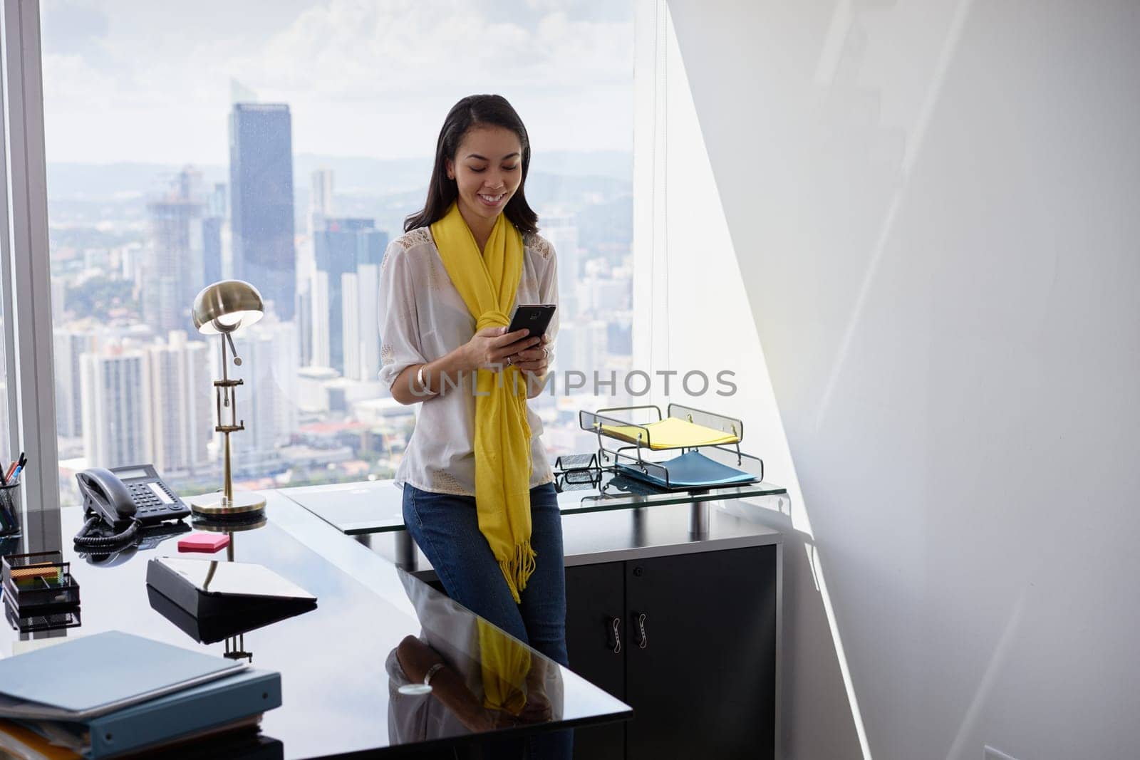 Young multiethnic Chinese Hispanic woman leaning on table in a modern office building, with a beautiful sight of the city in background. She holds a mobile phone and smiles happy