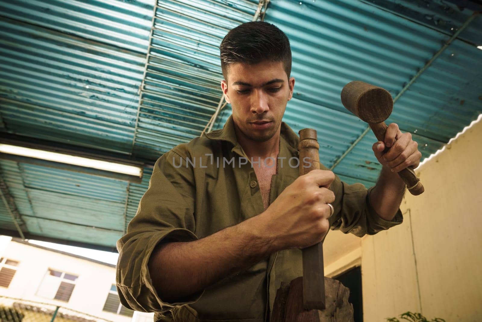 Young man at work learning craftsman profession, working with hammer and chisel. The artist carves a raw block of wood to make a wooden statue