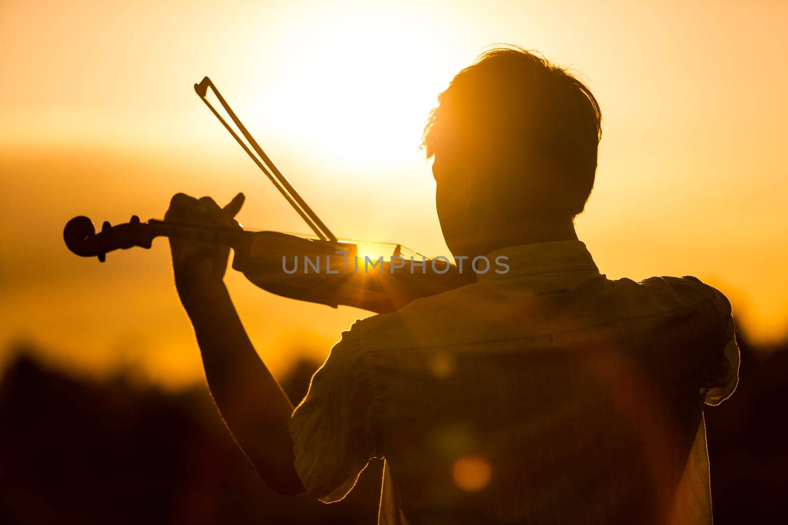 Young man or boy playing the violin at sunset standing with his back to the camera with his instrument and bow silhouetted against the fiery orange sky