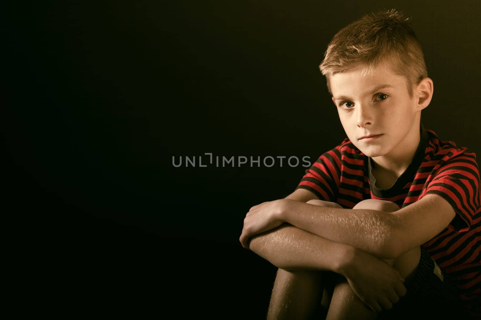 Close up Serious Boy Hugging his Knees and Looking Straight at the Camera Against Dark Green with Copy Space.