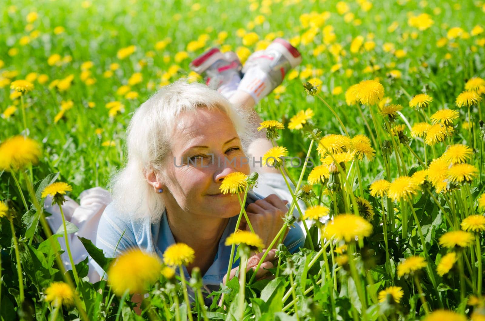 A woman lies in a clearing among the dandelions and sniffs a flower