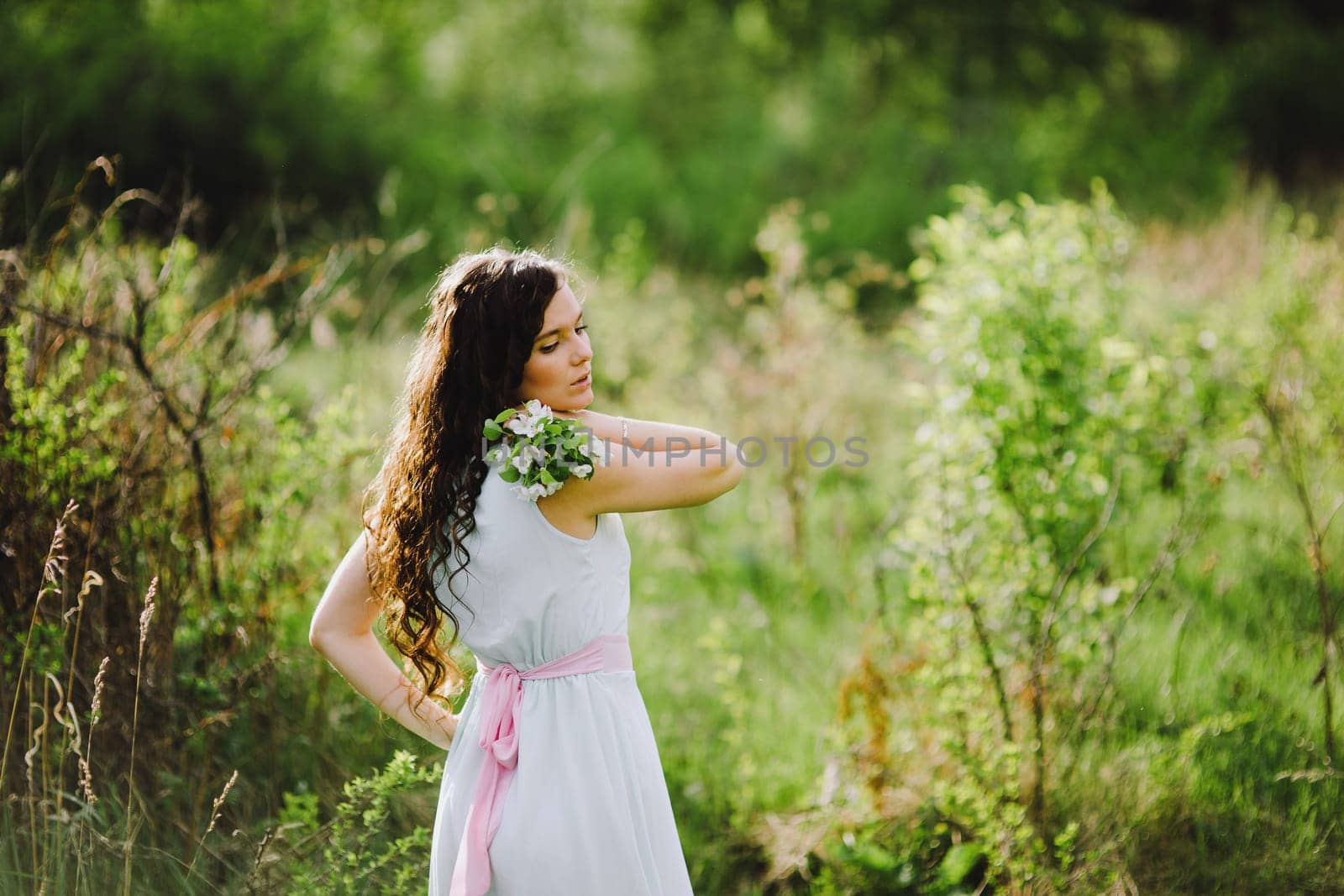 the girl with a bouquet of white flowers in a blue dress with a pink belt poses in the wood
