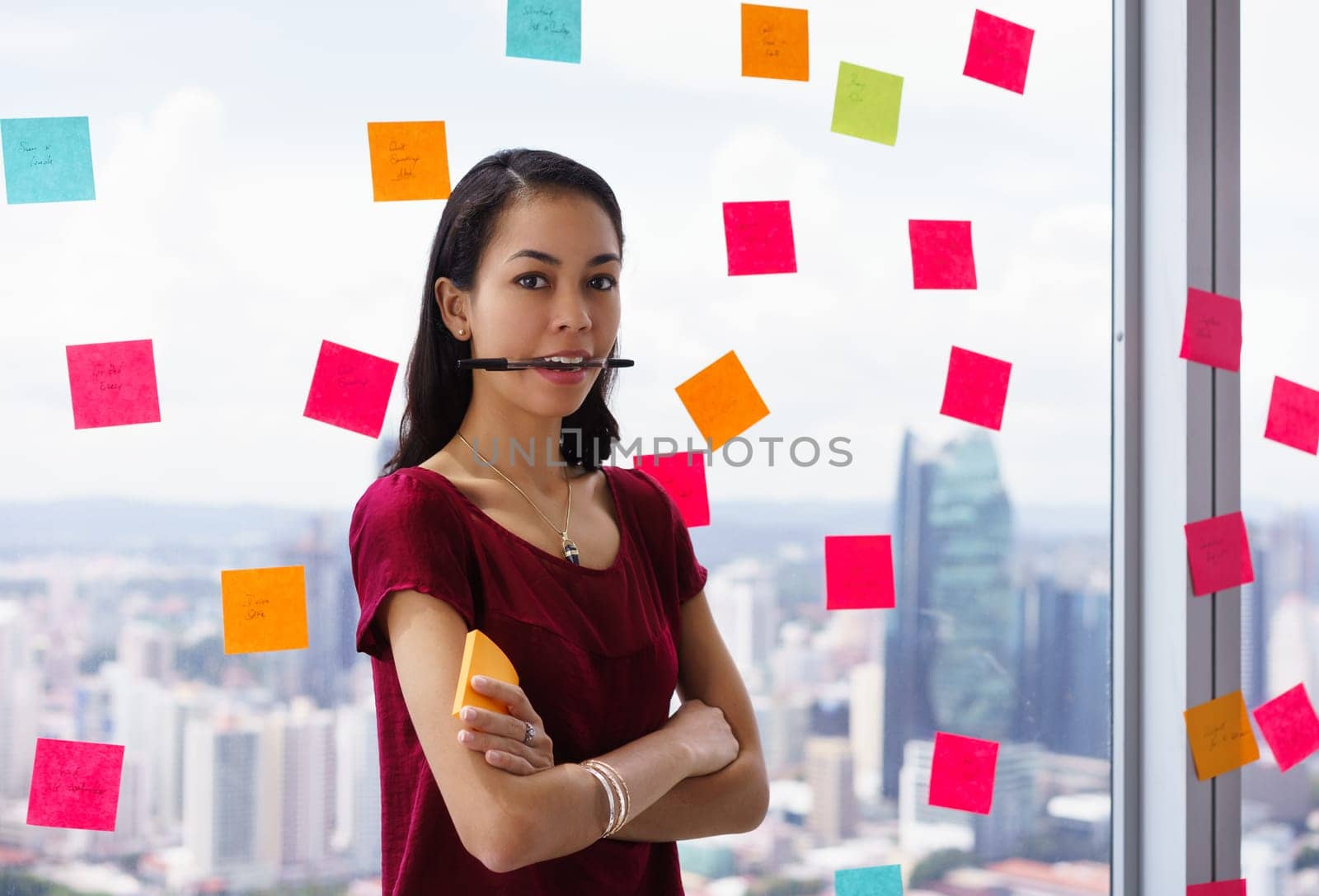 Portrait of proud assistant with pen in mouth. The mixed race woman stands in modern office against a window full of adhesive notes with tasks