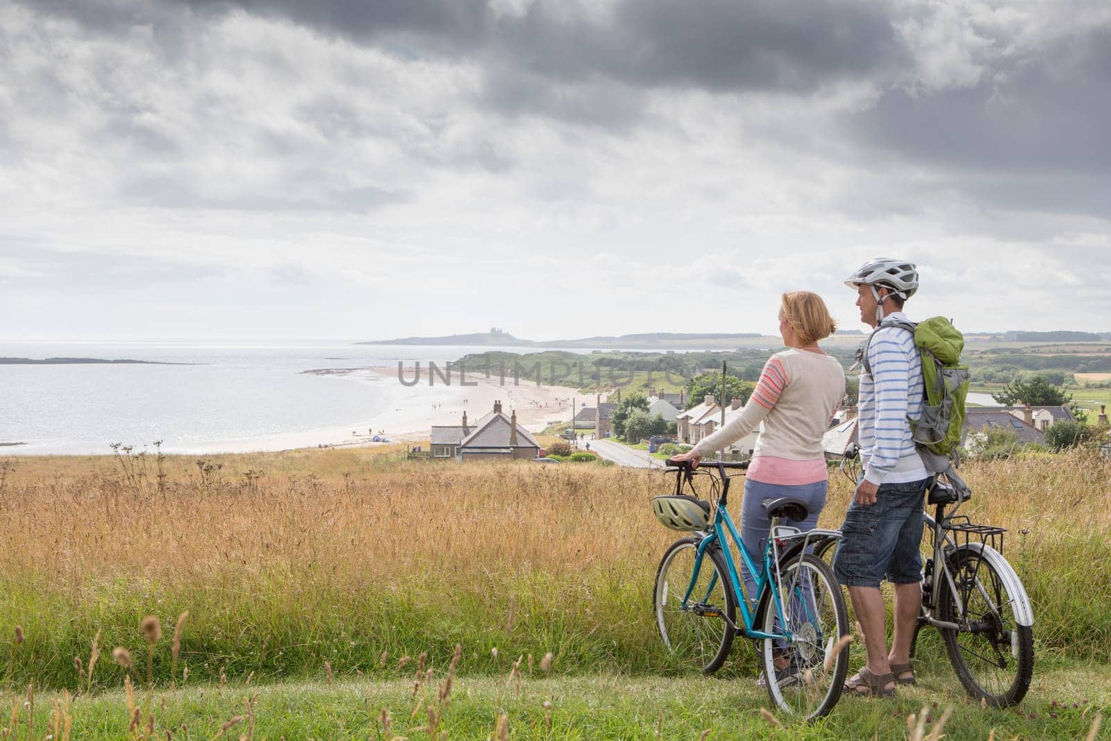 A couple are enjoying the view on the sand dunes. They are holding their bikes, looking at the beach.