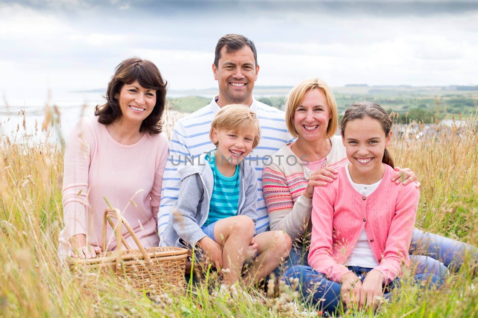 A family are sat on the beach with a picnic basket. They are all sitting down smiling, looking at the camera.