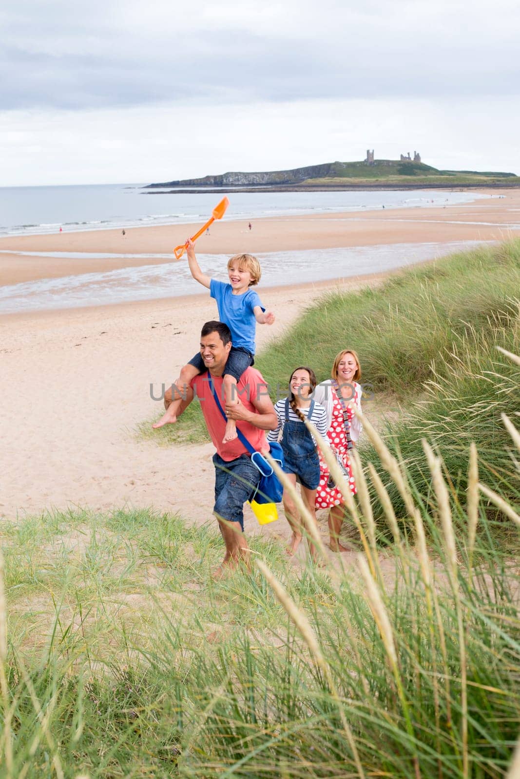 A family of four are walking up the sand dunes toegther, leaving the beach. The little boy is on his dads shoulders laughing.