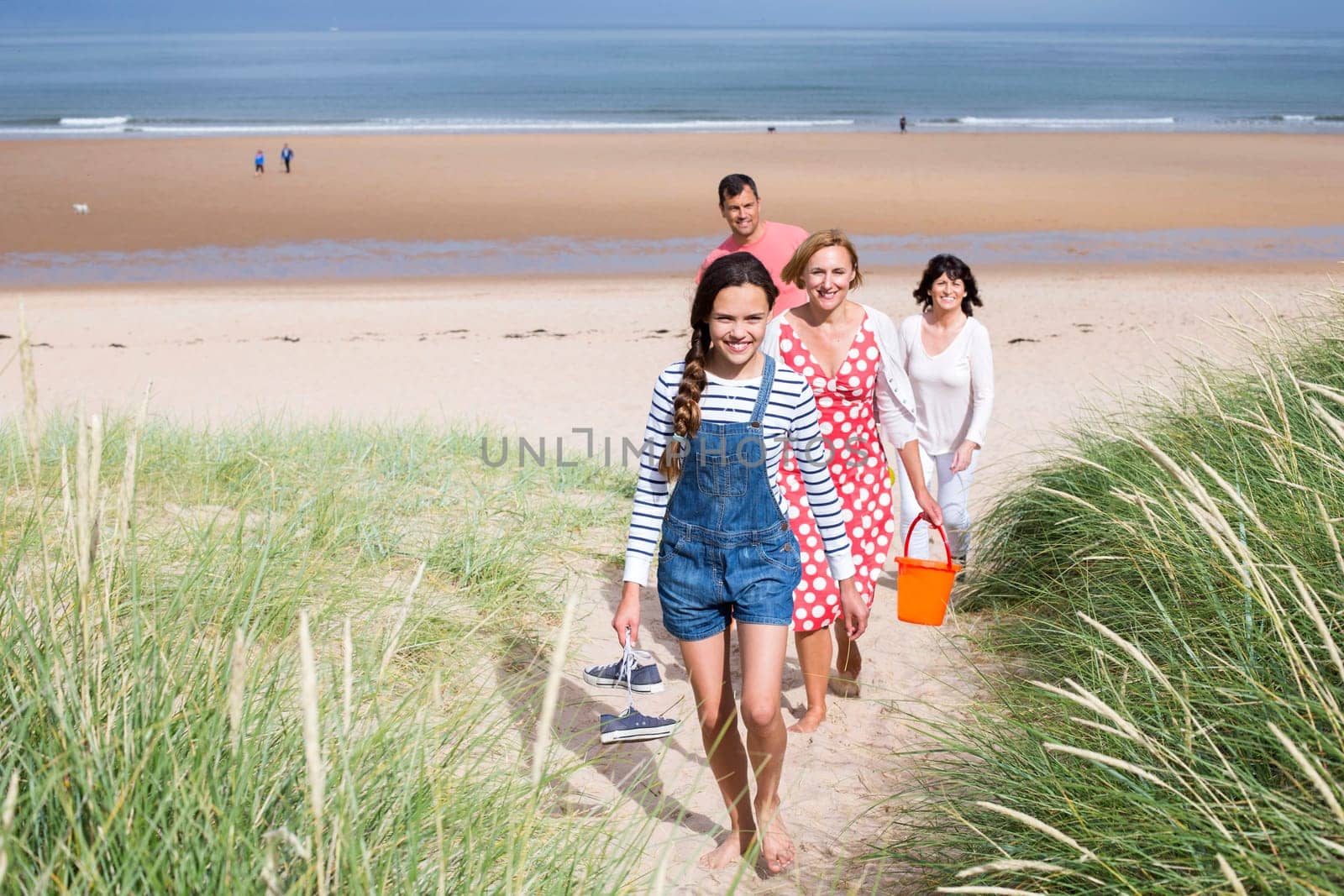 A family of four are walking up the sand dunes leaving the beach. They all look happy and are smiling.