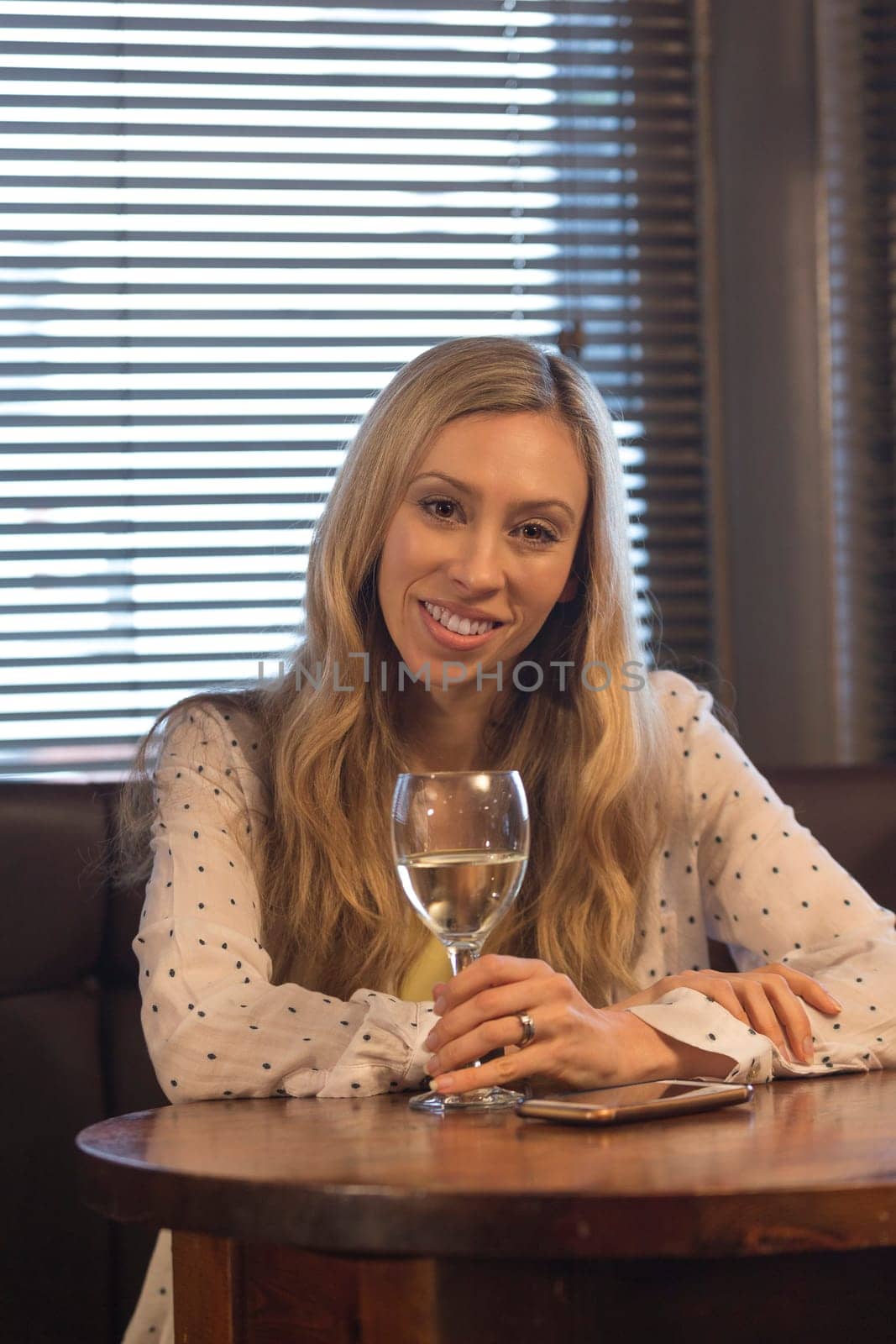 Attractive young woman sitting in a pub with a glass of wine and her smartphone. She is smiling at the camera.