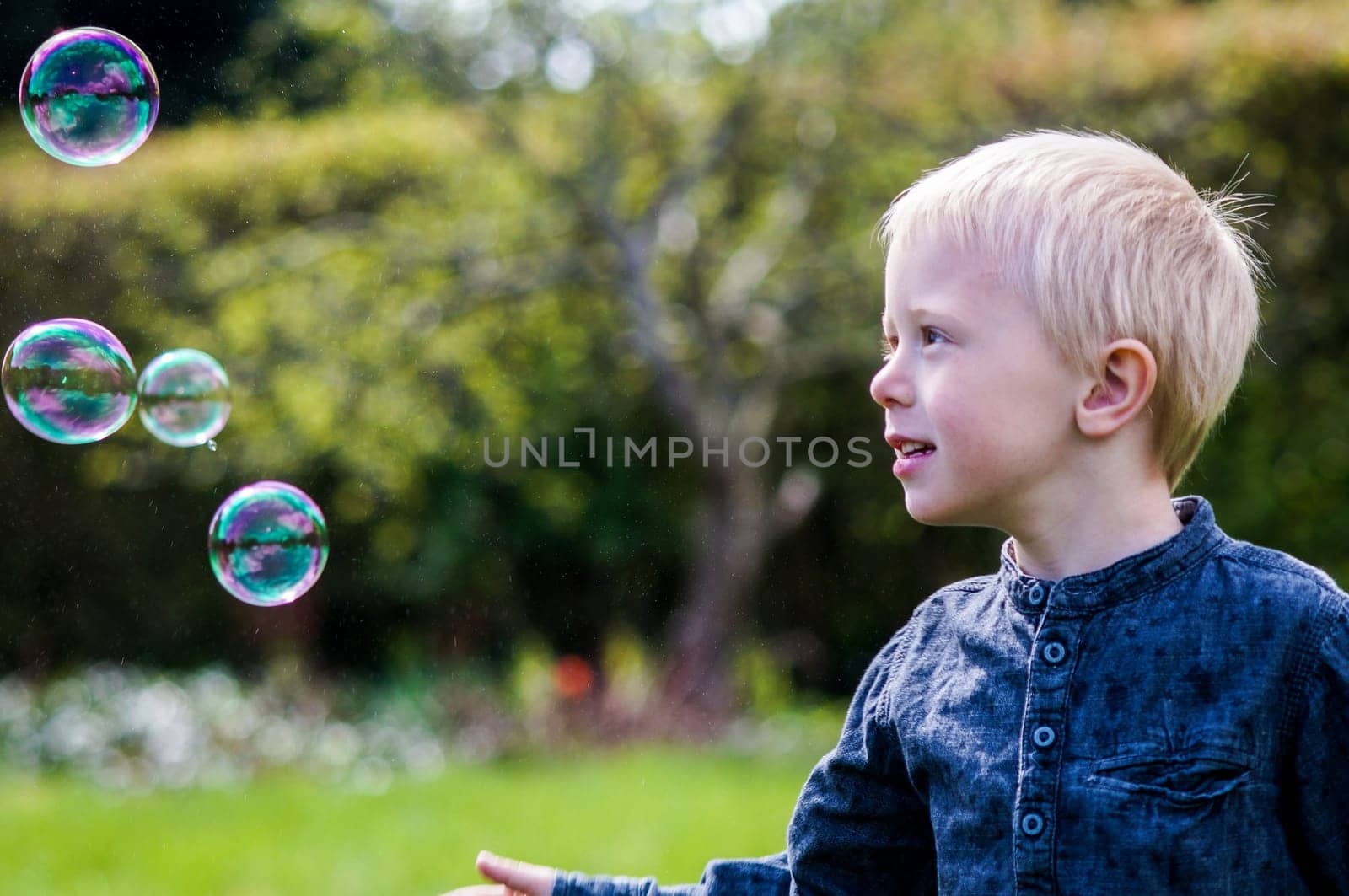 A Little boy blows soap bubbles in the garden on a summer day. He wears a blue shirt and is happy