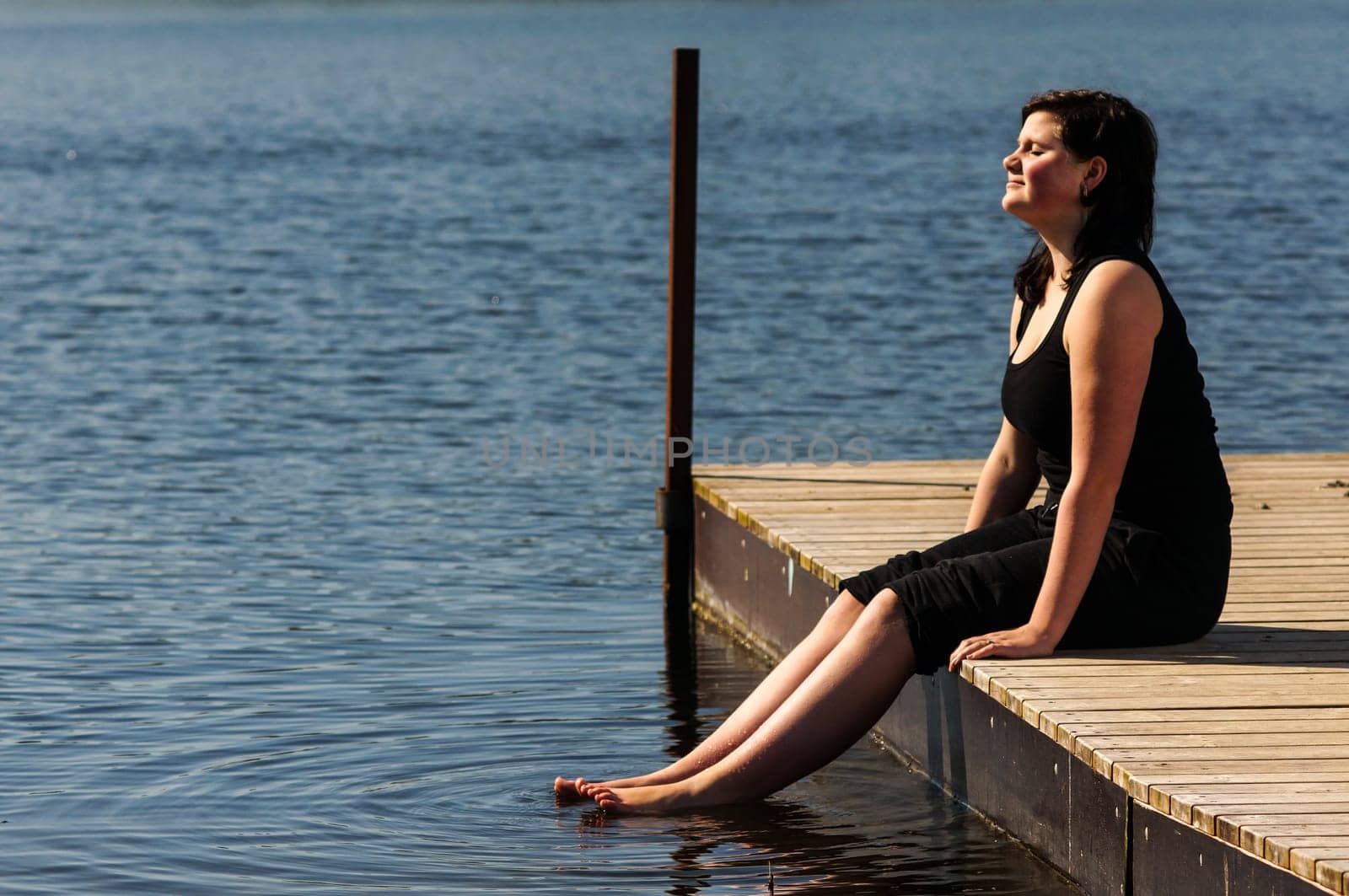 young lady sitting on the jetty and dips her feet in the cool lake water on a sunny day