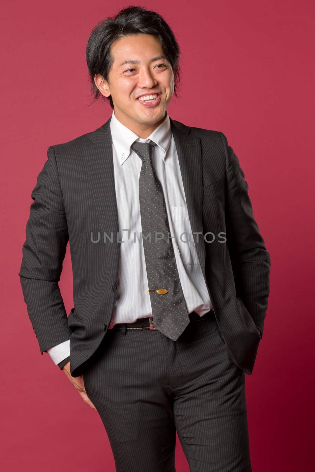 A half body portrait of a young Japanese man in a business suit laughing on a red background.