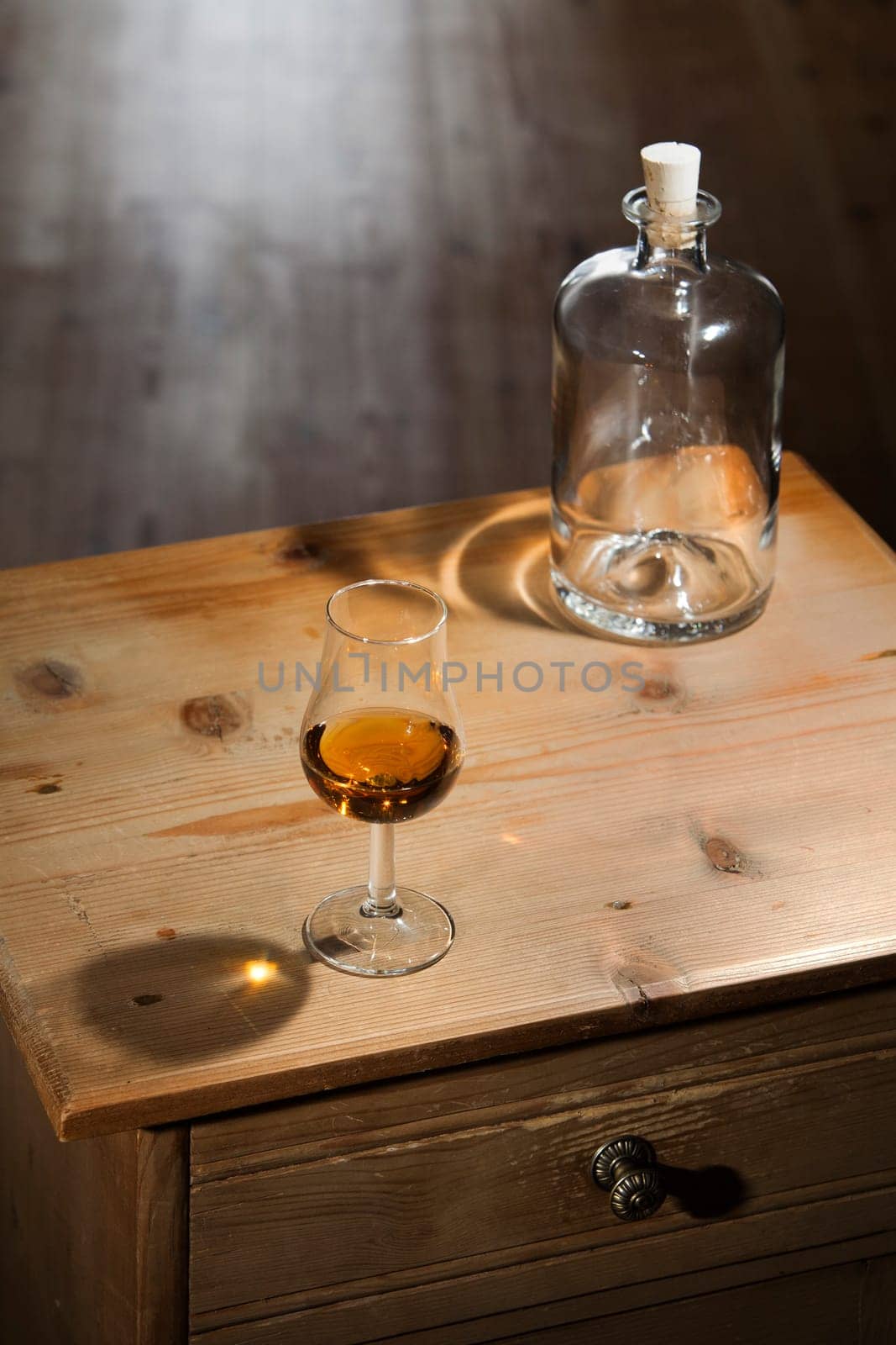 Empty bottle and a glass of brandy on a vintage wooden table with closed drawer