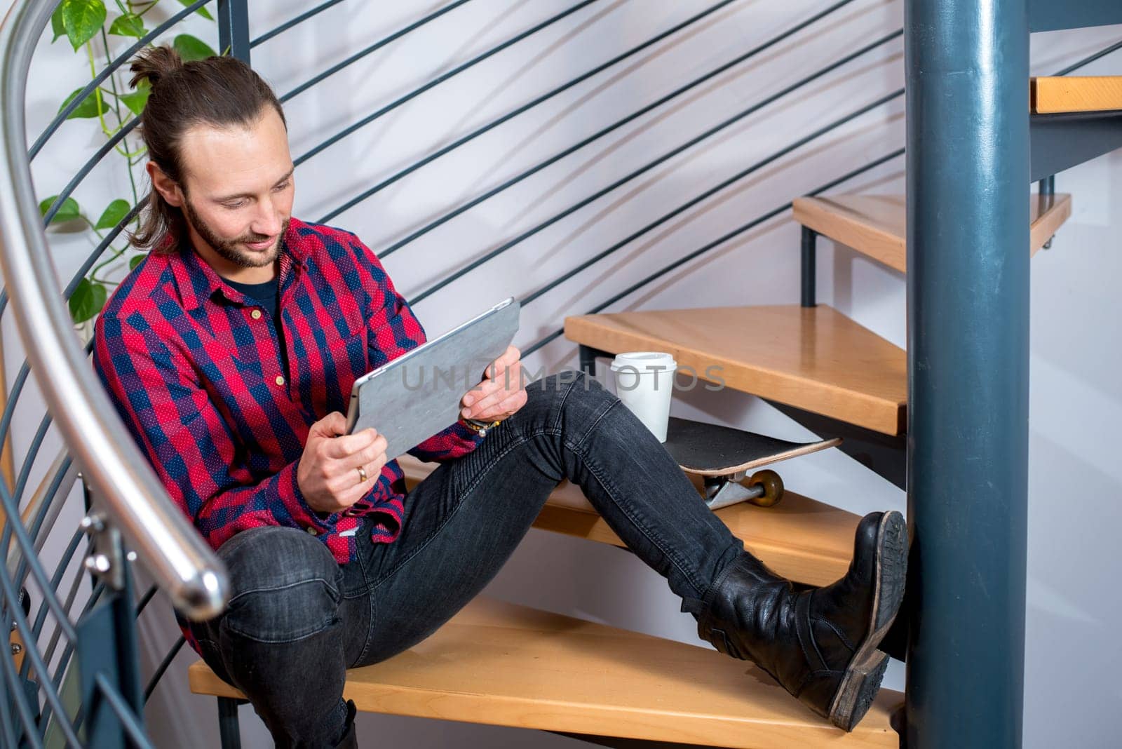 young bearted man in checkered shirt sitting on stairs and using tablet PC