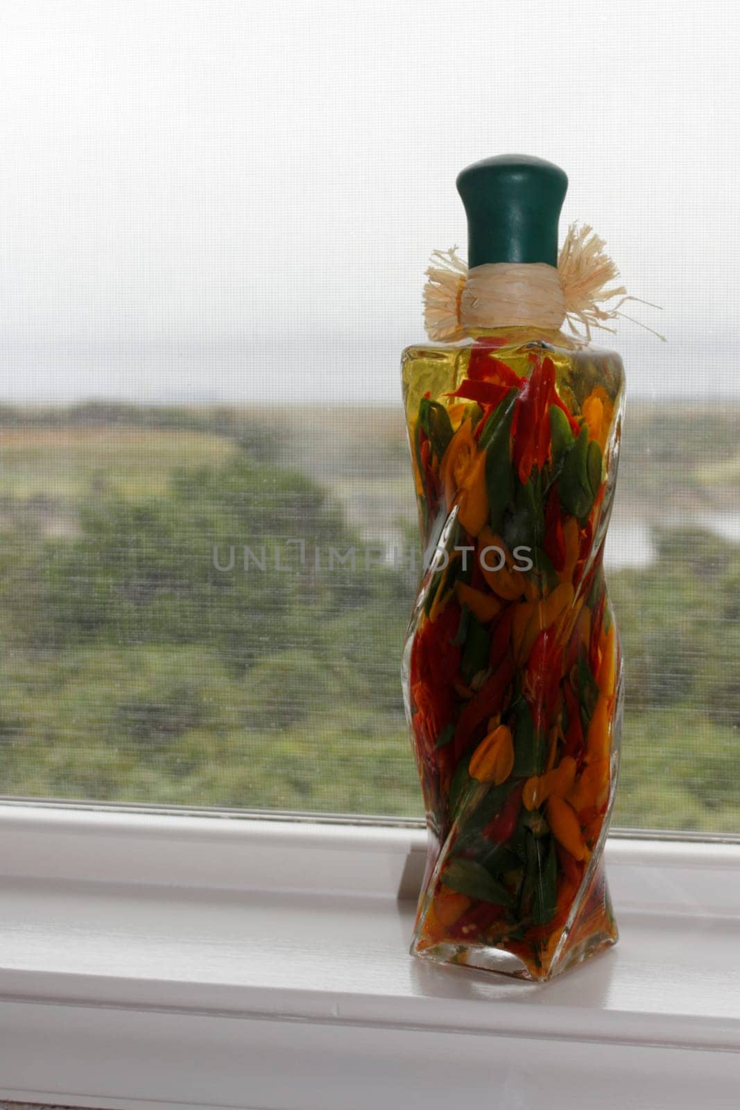 Tall glass bottle filled with vinegar and red green and yellow sweet bell peppers. Decorative bottle on a window sill with three colors of peppers and vinegar.