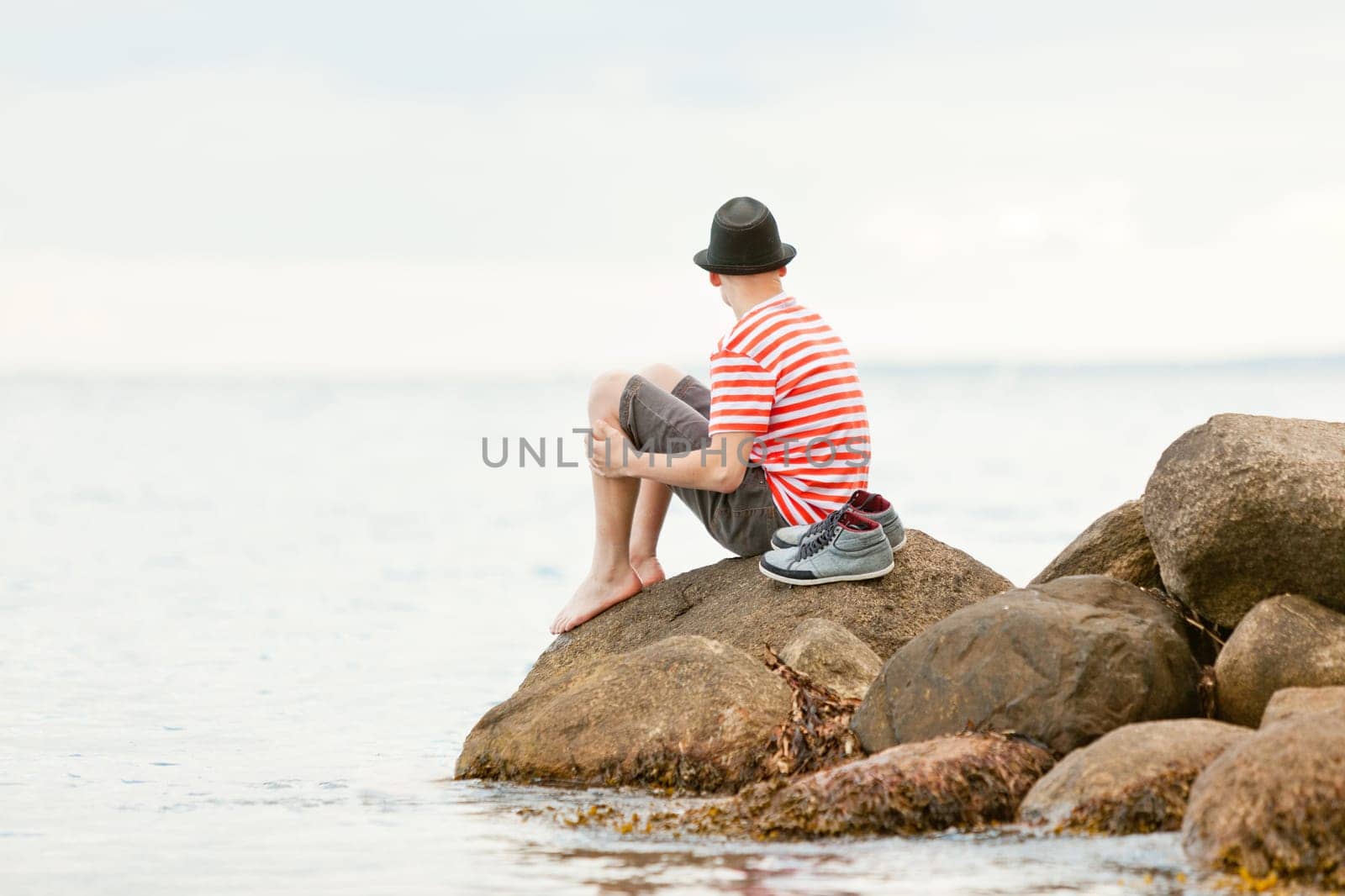Trendy barefoot young man sitting on a rock at the edge of the ocean staring away from the camera out over the water, with copy space